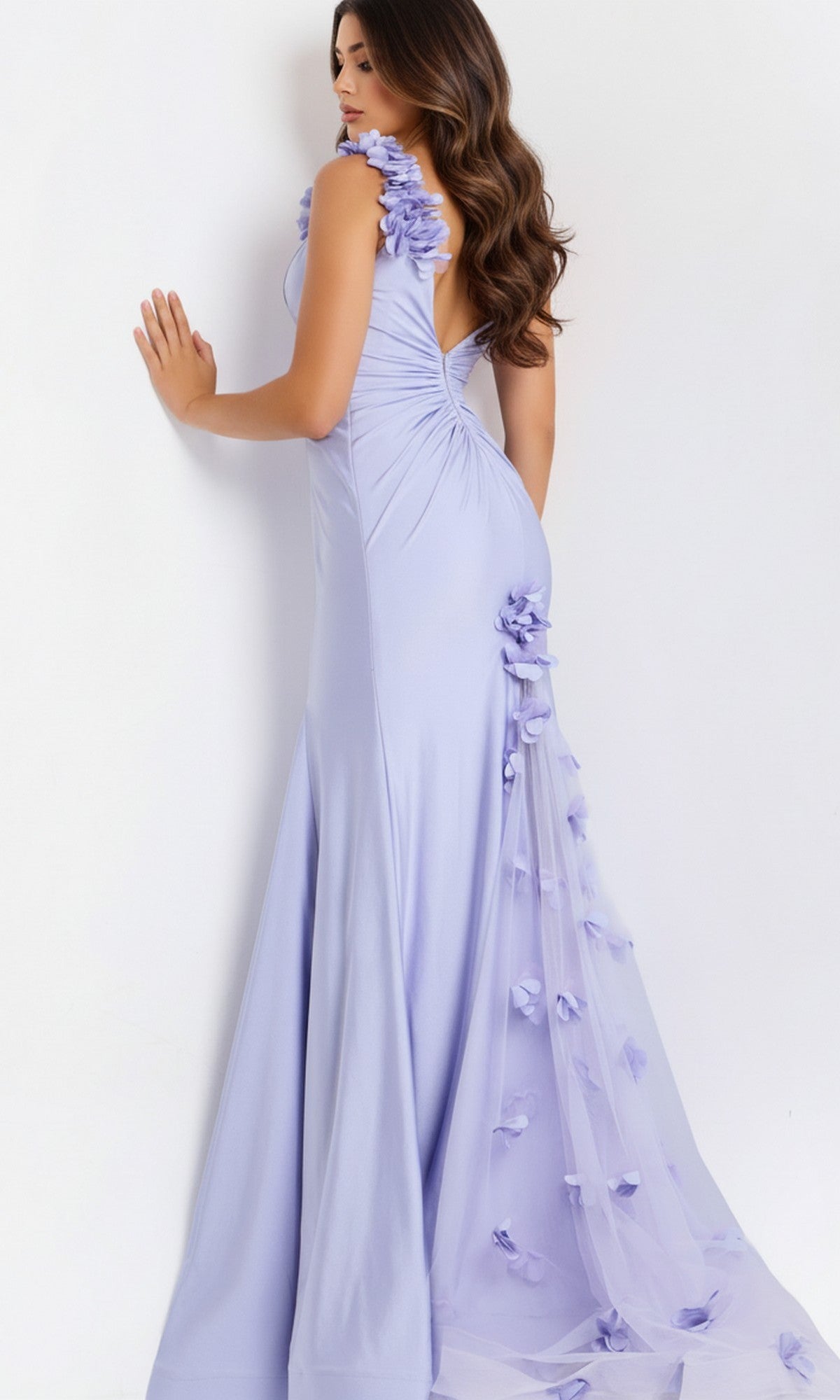 A woman wearing the Jovani 48120 long prom dress, featuring a light purple mermaid silhouette with 3D floral appliques and a tulle train, stands facing a white wall and looks to her left.